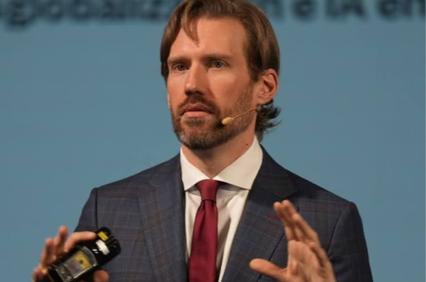 A man in a suit is speaking at a conference while holding a small device.