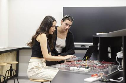 Two women are collaborating on a laptop in a modern workspace filled with electronic components.