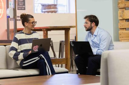 Two people are sitting on a couch, engaged in a conversation while using their laptops.
