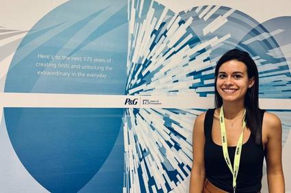 A woman smiling in front of a corporate anniversary banner that reads 'Here's to the next 175 years of creating firsts and unlocking the extraordinary in the everyday.'