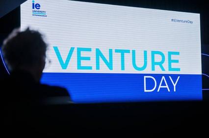 A person attends a Venture Day event at IE University, shown on a large presentation screen.