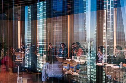 A group of professionals in a meeting seen through a reflective glass window overlaid with urban building reflections.