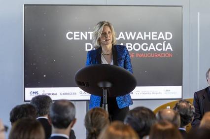 A woman delivers a speech at a podium with a backdrop displaying event logos and text.