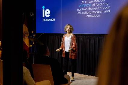 A woman presents at a conference in front of a banner that reads 'IE Foundation' with audience members in the foreground.