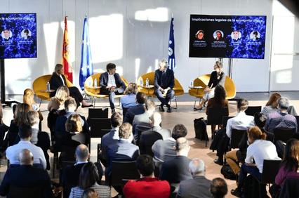 A conference setting with five speakers seated on stage in discussion in front of an audience.