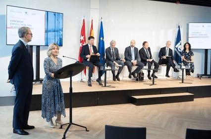 A woman is speaking at a podium in a conference room with several attendees seated behind her under national flags.
