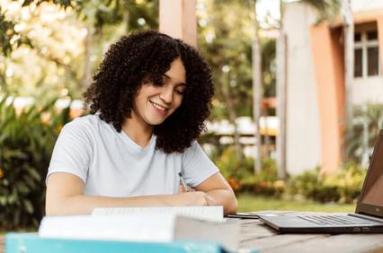 A young woman with curly hair smiling while studying outdoors with a laptop and books on a table.