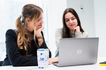 Two women are sitting at a table, engaged in a conversation while looking at a laptop.