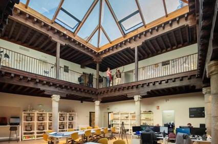 A spacious interior with a glass ceiling and wooden beams, featuring desks and people interacting.