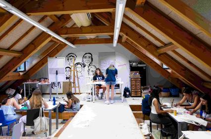 A creative workshop takes place in an attic, with several participants engaged in artistic activities.