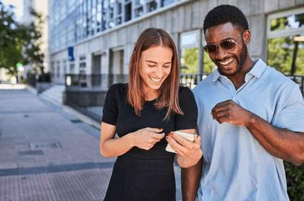 A man and a woman are smiling and looking at a smartphone together on a sunny city street.