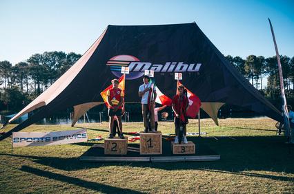Three athletes stand on a podium at an outdoor sporting event under a large tent with the Malibu logo.