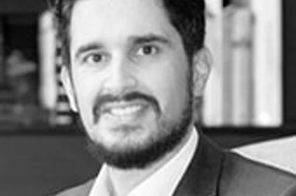 A professional black and white portrait of a smiling man with a beard, dressed in a suit and tie, standing in front of bookshelves.