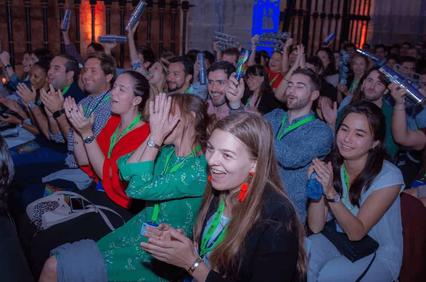 A group of people clapping and smiling during an event indoors, illuminated by blue lighting.