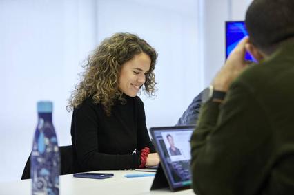 A woman with curly hair is smiling while using a laptop at a table in a bright environment.