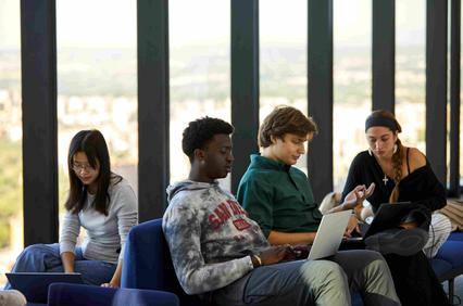 A group of young people working collaboratively on laptops in a modern space with large windows.