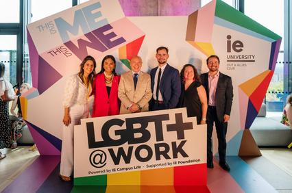 Group of six people smiling at a promotional event for LGBT@Work, standing behind a sign supported by IE University.