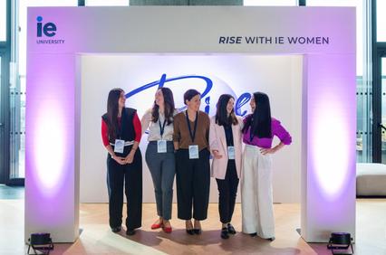Five women standing and interacting under a lit archway with the logo of IE University.