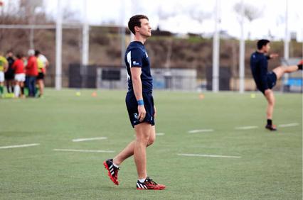 A young male athlete in a blue uniform standing on a soccer field, looking to the side, with other players training in the background.