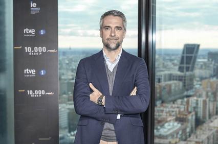 Man in a suit standing with arms crossed in front of a glass window overlooking a city.