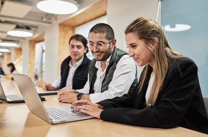 A group of three young professionals collaborating on a laptop in a modern office environment.