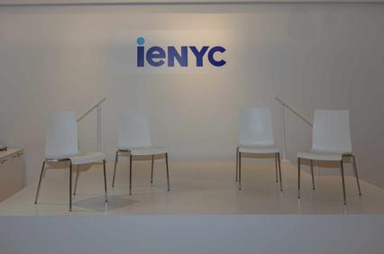 A minimalist room with four white chairs in front of a wall bearing the logo 'ieNYC'.