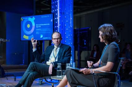 A man and a woman are engaged in a discussion during a panel at a conference, highlighted by modern lighting with an infographic presentation in the background.