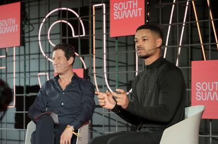 Two men are sitting on a stage, engaged in a discussion, with a neon 'South Summit' sign in the background.