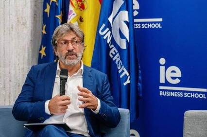 A man in a blue shirt and blazer speaks while holding a microphone, seated in front of banners of the EU and IE University Business School.