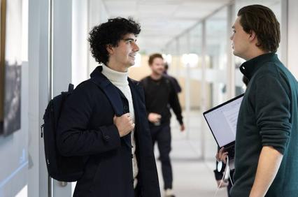 Two men talking in a corridor with one holding a backpack and the other holding documents.