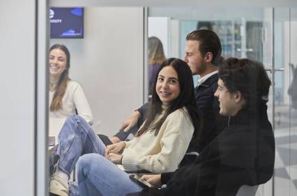 Four young adults are seen sitting and chatting casually inside a modern office environment.
