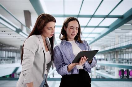 Two women are collaborating and discussing something on a tablet in a modern office setting.