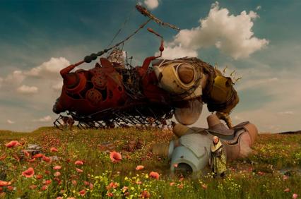 A surreal landscape featuring a large, abstract mechanical creature surrounded by vibrant flowers under a cloudy sky.