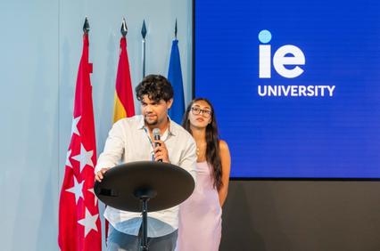 A young man speaks at a podium while a woman stands beside him during an event at IE University.