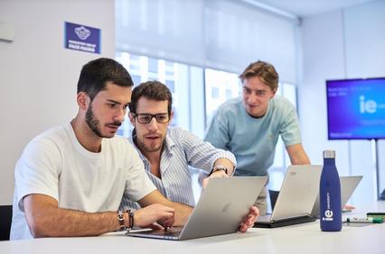 Three young men collaborate over a laptop in a modern office setting.