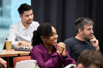 A group of people sitting at a table in a meeting, one woman is laughing while others are engaged in conversation.