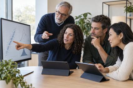 A group of four professionals discusses data displayed on a screen while using tablets.