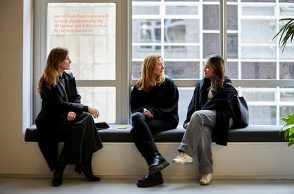 Three young women are sitting together on a bench, engaged in conversation near a large window.