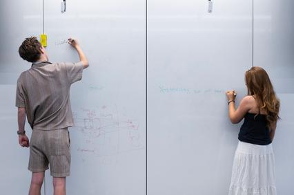 Two individuals are writing on a glass whiteboard in a modern workspace.