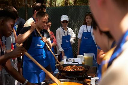 People in blue aprons are cooking and stirring a large paella outdoors.