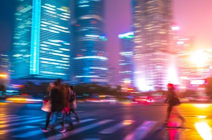 Blurry image of people walking across a crosswalk at night with illuminated skyscrapers in the background.