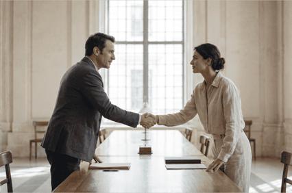 A man and a woman shake hands across a wooden table in a well-lit room.