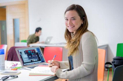 A young woman smiling at the camera while working on a laptop in a colorful study area.