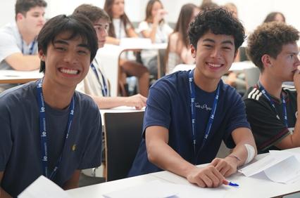 Two smiling young men are seated in a classroom with other students in the background.