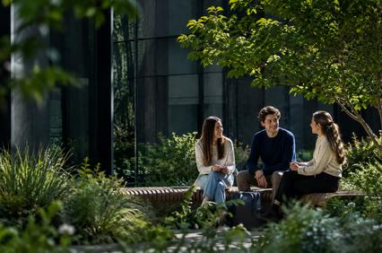 Three young people are sitting together in a green outdoor space, engaging in conversation.