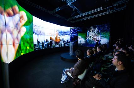 A large, modern presentation space with an immersive screen displaying various visuals and an audience seated in front.