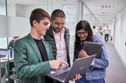 Three professionals, two men and one woman, are discussing over a laptop in a modern office corridor.