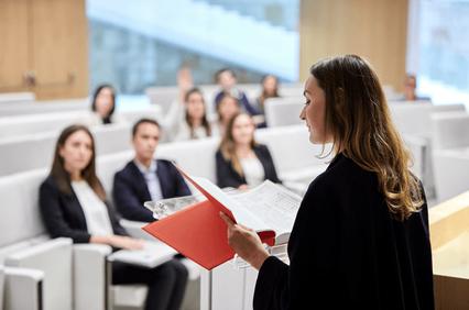 A woman stands presenting at a podium in a lecture hall filled with attendees.