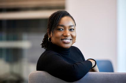 A smiling woman in a black turtleneck sitting on a gray sofa.