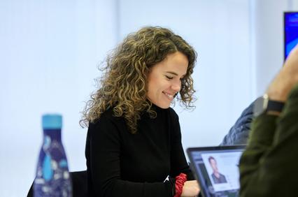 A woman with curly hair smiles while working at a desk with a laptop.